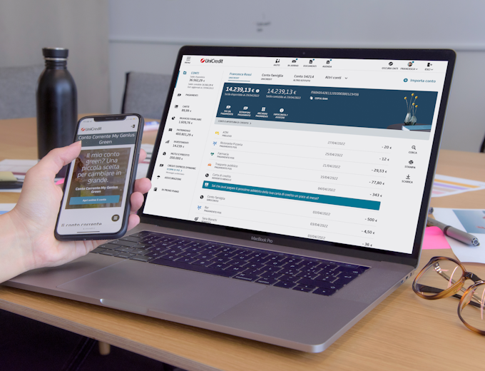 Hand with a mobile device in front of the laptop, both showing UniCredit interface