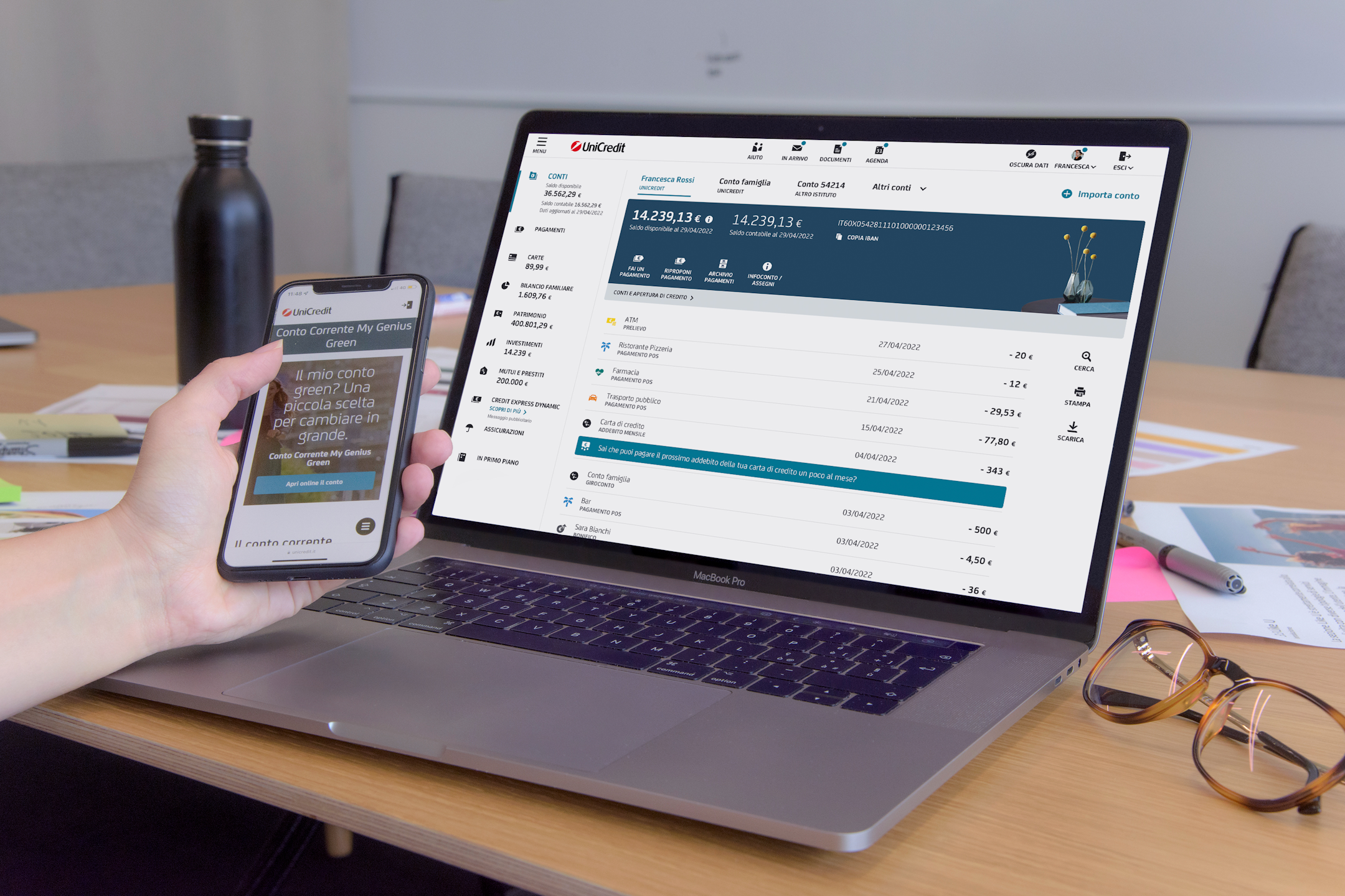 Hand with a mobile device in front of the laptop, both showing UniCredit interface