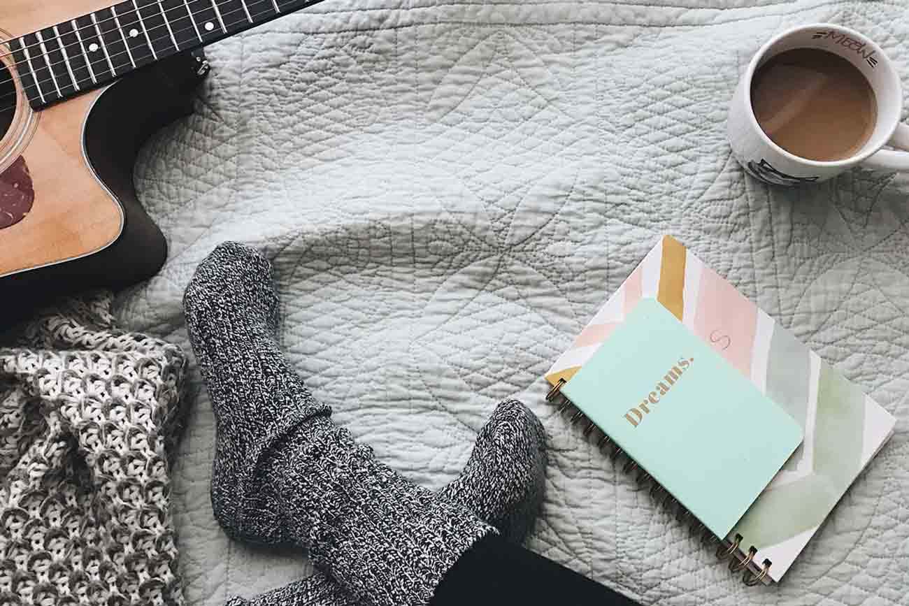Woman sitting on bed with guitar, coffee, and notebook