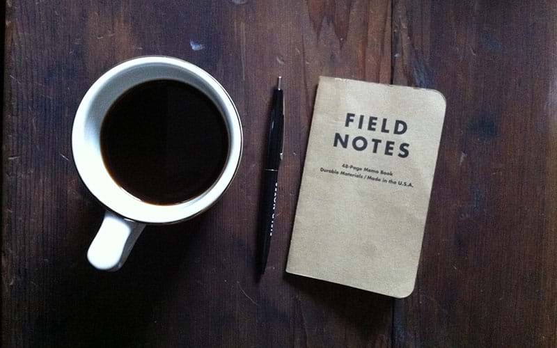 Cup of coffee on table next to booklet that says field notes