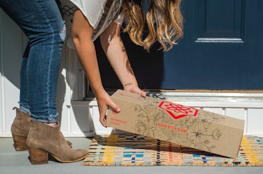 Person in brown ankle boots and jeans picking up a floral-decorated delivery box off a welcome mat in front of a navy blue door on a sunny day.