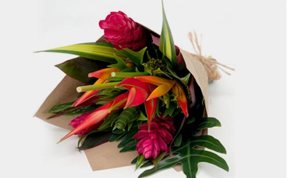 Vibrant bouquet of tropical flowers wrapped in brown paper, featuring red ginger, heliconia, and lush green foliage on a white background.