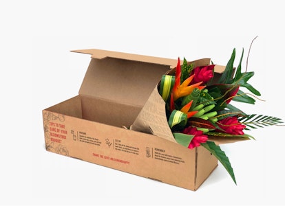 Colorful bouquet of fresh flowers including red, orange and yellow blooms with green foliage, neatly arranged in an open cardboard box on a white background.