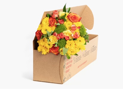 Vibrant bouquet of flowers including yellow blossoms and red roses arranged in a cardboard box against a neutral background, symbolizing a fresh floral delivery.
