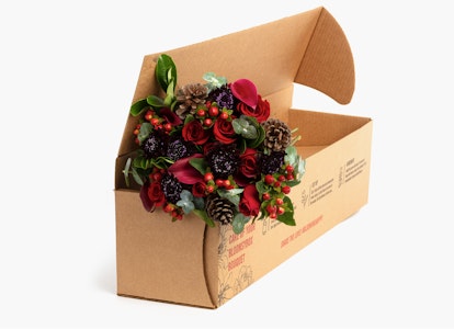 A festive holiday wreath with greenery, pine cones, and red berries partially nestled in an open cardboard box, against a clean, neutral background.