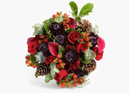 Beautiful floral arrangement with vibrant red roses, pink calla lilies, dark purple accents, pine cones, and green foliage on a white background.