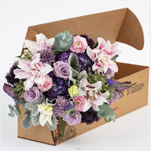 A vibrant bouquet of flowers with a mix of pink roses, purple blooms, and delicate white flowers, nestled in a brown cardboard delivery box against a white background.