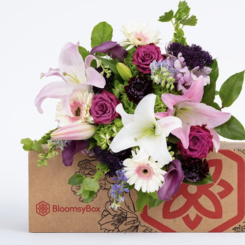 A vibrant BloomsyBox with an assortment of fresh flowers, including pink roses, white lilies, and purple accents, displayed against a white background.