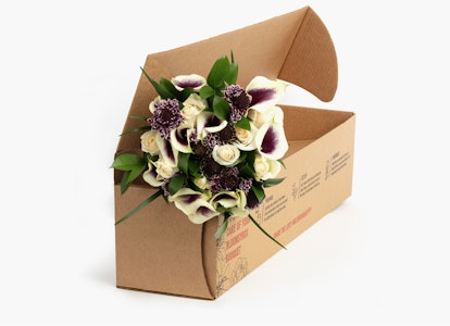 Bouquet of purple and white flowers with green leaves presented in a diagonal craft paper box on a clean, white background, suitable for gifting or decoration.