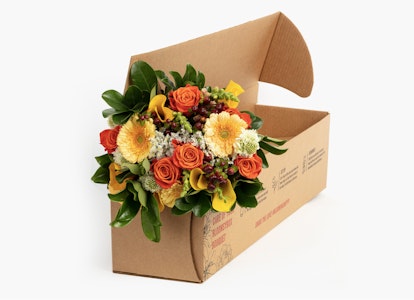 A vibrant bouquet of orange and yellow flowers with green leaves arranged beautifully in a brown cardboard delivery box against a white background.