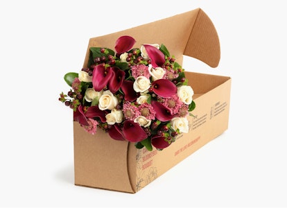 A vibrant bouquet of fresh flowers with roses and greenery elegantly arranged and presented in an open cardboard box against a neutral background.