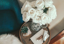 Elegant white roses arranged in a patterned vase on a round side table, with a cozy blue armchair, a small succulent, and skincare products, creating a serene home decor vignette.