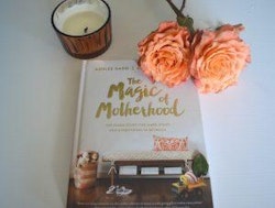 A cozy reading setup with a book titled 'The Magic of Motherhood,' fresh orange roses, and a lit scented candle on a white surface.