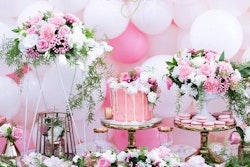 Elegant party table setup with pink theme, featuring a decorative cake with dripped icing, macarons, large balloons, and lush floral arrangements.