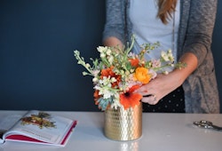 Woman in a grey cardigan arranging a bouquet of colorful flowers in a gold vase on a table with a floral arrangement book and scissors beside it.