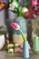 Vibrant pink tulip elegantly displayed in a small blue polka-dotted vase with a soft-focus background of assorted flowers and elegant decor on a wooden surface.