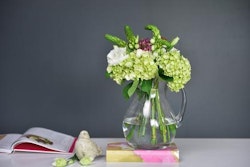 Vibrant bouquet of green hydrangeas and pink wildflowers in a clear glass vase on a table with books and a decorative bird, against a grey background.