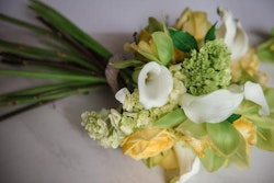 Elegant bridal bouquet featuring white calla lilies, yellow roses, and green hydrangeas with long stems tied together against a soft neutral background.
