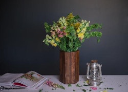 A rustic wooden vase with an arrangement of fresh yellow and green flowers next to a glass lantern and an open book on a table with scattered petals.