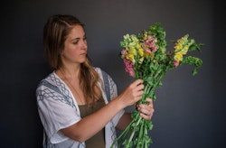 A woman in a white and blue blouse thoughtfully arranges a colorful bouquet of snapdragons, showcasing shades of pink, yellow, and green, against a serene gray background.