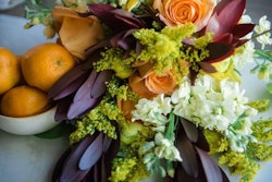 Elegant floral arrangement featuring orange roses, deep purple leaves, and accents of yellow and white flowers, artfully displayed beside fresh oranges on a table.