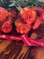 Bunch of vibrant orange and yellow roses with green leaves lying on a wooden table, featuring a red ribbon with the BloomsyBox logo printed in white.