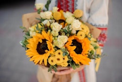 Close-up of a person holding a vibrant bouquet featuring sunflowers, white roses, and other mixed flowers wrapped in brown paper, embodying a warm, rustic aesthetic.