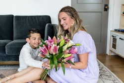 A smiling woman in a blue dress receives a large bouquet of pink lilies from a young boy in a gray sweater, both sitting on a wooden floor in a cozy living room.