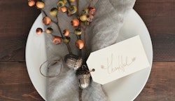 Elegant Thanksgiving place setting with a white plate, decorative autumnal sprig, and a name card with handwritten "thankful" on a rustic wooden table.