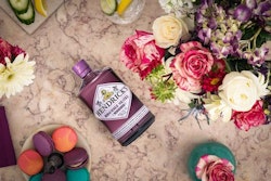 Top-down view of a Hendrick's gin bottle surrounded by vibrant flowers, colorful macarons, and sliced citrus fruits on a marble table, conveying a luxurious and festive ambiance.