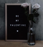 A romantic Valentine's Day setup featuring a black felt letter board with the message "BE MY VALENTINE" in white letters, accompanied by two pale pink roses in a clear glass vase.