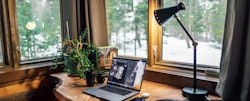 Cozy home office setup with a laptop open on Adobe Photoshop, a desk lamp, and a plant beside a window overlooking a snowy forest.