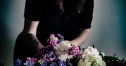 A person in a dark t-shirt arranging a vibrant bouquet of flowers, including purple, blue, and white blooms, with a soft-focus background.