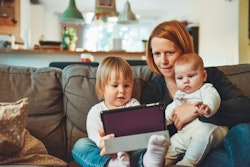 A mother sits on a couch cuddling her two young children, a toddler and a baby, as they look at a tablet together in a cozy, well-lit living room.