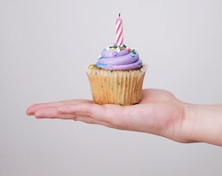 Hand holding a birthday cupcake with purple frosting and a lit candle against a neutral background, conveying a celebration or a birthday party theme.