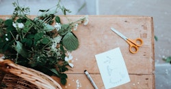 Rustic wooden table with a bouquet of wildflowers, orange scissors, a pen, and a sketch of a flower on a card, next to a woven basket.