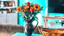 A vibrant bouquet of sunflowers and roses in a black vase on a teal tablecloth, with a blurry television and cozy living room setting in the background.