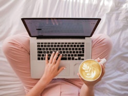Person sitting on a bed with pink bedding, working on a laptop, holding a cup of coffee with latte art visible. The focus is on comfort and productivity at home.