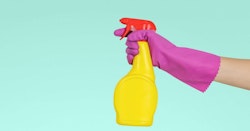 A person wearing a pink rubber glove holds a yellow spray bottle against a soft teal background, indicating house cleaning or disinfecting.