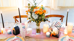 Elegant wedding table setting with orange blooms centerpiece, lit candles, delicate pink runner, and modern geometric accents in a luminous room with wooden chairs.