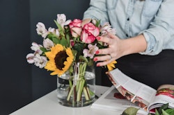 Person arranging a vibrant bouquet of sunflowers and pink roses in a clear vase on a table with a magazine in the background.