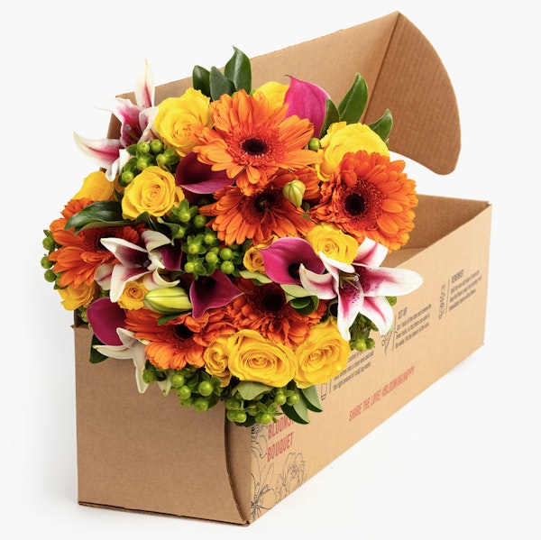 Vibrant bouquet of orange gerberas, yellow roses, and purple lilies with green accents, beautifully arranged and presented in a brown cardboard flower box.