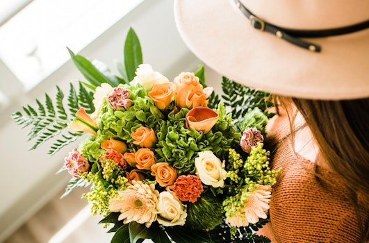 Close-up of a vibrant bouquet with orange and peach roses, green foliage, held by a person in a stylish hat, suggesting a sunny, fashionable setting.