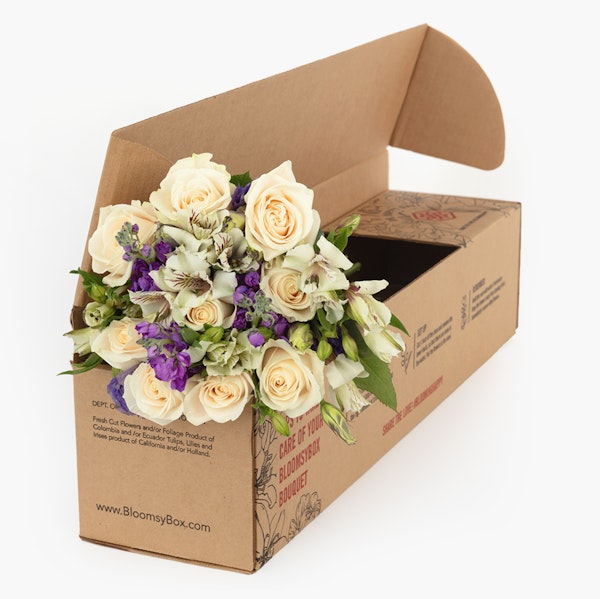 Bouquet of white roses and purple flowers packaged in a brown cardboard BloomsyBox, prepared for delivery against a white background.