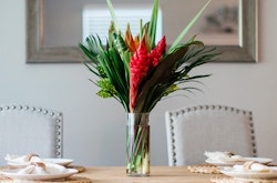 Vibrant tropical flower arrangement in a clear glass vase on a wooden dining table, flanked by elegant chairs and table settings in a well-lit room.