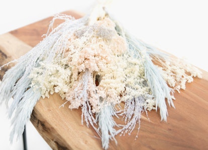A bouquet of dried flowers in pastel colors arranged on a diagonal wooden plank, set against a clean white background, evoking a rustic and delicate aesthetic.
