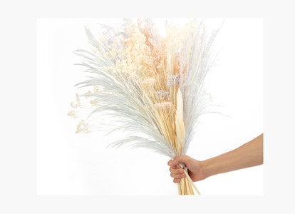 Hand holding a bouquet of dried flowers with various textures and colors including white, orange, and beige against a light background.