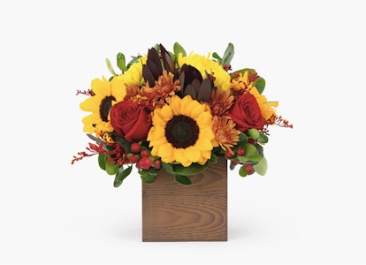 Vibrant floral arrangement featuring sunflowers, red roses, and yellow blooms with greenery in a wooden box, displayed against a white background.