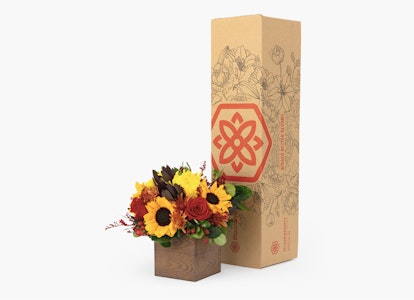 Elegant floral arrangement with sunflowers and autumn leaves beside a cardboard packaging box with ornate patterns and a red emblem on a white background.
