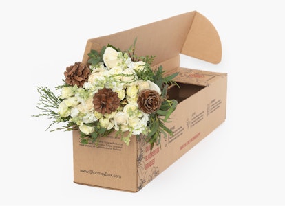 Elegant white flowers and green foliage arrangement with pine cones in a cardboard delivery box placed against a light gray background.